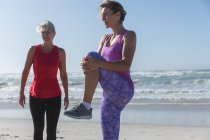 Due amiche caucasiche che si esercitano su una spiaggia in una giornata di sole, praticano yoga e si allungano con il mare sullo sfondo. — Foto stock