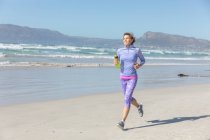 Кавказька жінка, яка в сонячний день насолоджується вправами на пляж, біжить на березі, посміхається і тримає пляшку води.. — стокове фото