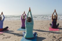Grupo de amigas caucásicas disfrutando haciendo ejercicio en una playa en un día soleado, practicando yoga y sentadas en posición de yoga. - foto de stock