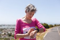 Senior Caucasian woman enjoying exercising on a sunny day, taking a break after running race, wearing numbers checking her smartwatch. — Stock Photo