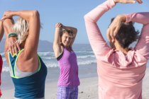 Grupo de amigas caucásicas disfrutando haciendo ejercicio en una playa en un día soleado, practicando yoga y estirándose con el mar al fondo. - foto de stock