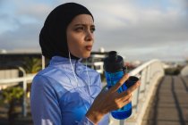 Fit gemischte Rennläuferin in Hijab und Sportbekleidung, die an einem sonnigen Tag im Freien in der Stadt trainiert und eine Wasserflasche in der Hand hält, die mit Kopfhörern auf einer Fußgängerbrücke Pause macht. Bewegung im städtischen Lebensstil. — Stockfoto