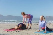 Groupe d'amies caucasiennes profitant de l'exercice sur une plage par une journée ensoleillée, pratiquant le yoga et debout et assis en position de yoga. — Photo de stock