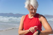 Mulher caucasiana sênior gosta de se exercitar em uma praia em um dia ensolarado, de pé e usando seu smartwatch com o mar no fundo. — Fotografia de Stock