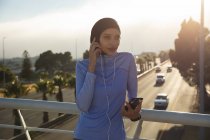 Fit gemischte Rennläuferin in Hijab und Sportbekleidung, die an einem sonnigen Tag im Freien in der Stadt trainiert und während des Trainings mit Smartphone und Kopfhörer auf einer Fußgängerbrücke eine Pause einlegt. Bewegung im städtischen Lebensstil. — Stockfoto