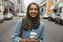 Ritratto di donna di razza mista che indossa hijab fuori e in giro per la città, in piedi in strada tenendo caffè da asporto, sorridendo alla telecamera. Stile di vita moderno pendolare. — Foto stock