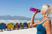 Donna caucasica anziana che si diverte ad allenarsi su una spiaggia in una giornata di sole, riposandosi dopo aver corso sulla riva del mare e bevendo acqua da una bottiglia con piccole case colorate sullo sfondo. — Foto stock