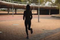 Vista trasera de la mujer de raza mixta en forma con hijab y ropa deportiva ejerciendo al aire libre en la ciudad, corriendo en un parque urbano. Ejercicio urbano. - foto de stock