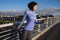 Fit mixed race woman wearing hijab and sportswear exercising outdoors in the city on a sunny day, stretching during workout using smartphone and earphones on a footbridge. Urban lifestyle exercise. — Stock Photo