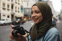 Mixed race woman wearing hijab sightseeing out and about on the go in the city, smiling holding digital camera taking photos. Tourism sightseeing modern lifestyle. — Stock Photo