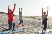 Group of Caucasian female friends enjoying exercising on a beach on a sunny day, practicing yoga and standing in yoga position. — Stock Photo