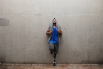 Uomo disabile di razza mista con una gamba protesica, che si allena in un parco urbano, indossa un top con cappuccio appoggiato a un muro che tiene la corda per saltare una pausa. Fitness disabilità stile di vita sano. — Foto stock