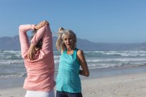 Due amiche caucasiche anziane che si esercitano su una spiaggia in una giornata di sole, praticano yoga e si allungano con il mare sullo sfondo. — Foto stock