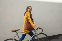 Mujer de raza mixta vistiendo hijab y jersey amarillo de ida y vuelta en la ciudad, sonriendo caminando con bicicleta. Commuter estilo de vida moderno. - foto de stock