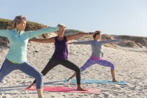 Groupe d'amies caucasiennes profitant de l'exercice sur une plage par une journée ensoleillée, pratiquant le yoga et se tenant debout en position de yoga. — Photo de stock