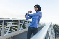 Fit gemischte Rennläuferin in Hijab und Sportbekleidung, die an einem sonnigen Tag im Freien in der Stadt trainiert und ihre Arme auf einer Fußgängerbrücke ausstreckt. Bewegung im städtischen Lebensstil. — Stockfoto