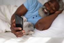 Hombre mayor afroamericano acostado en una cama en un dormitorio, usando un teléfono inteligente y sonriente, distanciamiento social y aislamiento en cuarentena - foto de stock