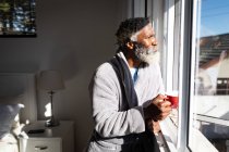 Ein älterer afroamerikanischer Mann steht in einem Schlafzimmer, hält eine Tasse Kaffee in der Hand, blickt durch ein Fenster, soziale Distanzierung und Selbstisolierung in Quarantäne — Stockfoto
