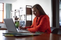 Mujer caucásica disfrutando del tiempo en casa, distanciamiento social y autoaislamiento en cuarentena, sentada en la mesa, usando una computadora portátil, tomando notas. - foto de stock