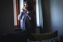 Кавказская женщина с длинными темными волосами наслаждается временем дома, социальным дистанцированием и самоизоляцией в карантинной изоляции, стоя, глядя в окно и держа чашку кофе. — стоковое фото