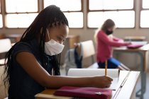 Due bambini multietnici seduti alle scrivanie con maschere facciali in classe. Istruzione primaria distanza sociale sicurezza sanitaria durante la pandemia di Covid19 Coronavirus — Foto stock