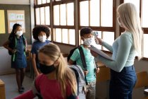 Professora branca com máscara facial medindo a temperatura de crianças em uma escola primária. Educação primária distanciamento social segurança sanitária durante Covid19 pandemia de coronavírus. — Fotografia de Stock