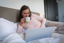 Mujer caucásica disfrutando del tiempo en casa, distanciamiento social y autoaislamiento en cuarentena, acostada en la cama en el dormitorio, usando una computadora portátil, bebiendo té. - foto de stock