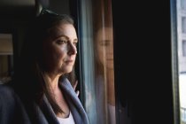 Donna caucasica con lunghi capelli scuri godendo del tempo a casa, distanza sociale e isolamento in quarantena, in piedi e guardando fuori dalla finestra. — Foto stock