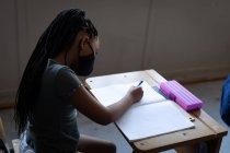 Дівчина змішаної раси носить маску для обличчя, сидячи на столі в школі. Початкова освіта соціальне дистанціювання охорона здоров'я під час пандемії коронавірусу Covid19 . — стокове фото