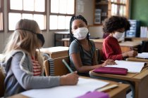 Gruppo multietnico di bambini delle elementari seduti alle scrivanie che indossano maschere facciali in classe. Istruzione primaria distanza sociale sicurezza sanitaria durante la pandemia di Covid19 Coronavirus. — Foto stock