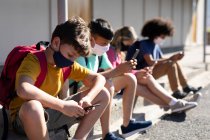 Multi ethnic group of elementary school kids wearing face masks using smartphones while sitting together. Primary education social distancing health safety during Covid19 Coronavirus pandemic — Stock Photo