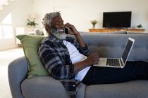 Hombre mayor afroamericano acostado en un sofá, usando una computadora portátil, hablando por teléfono, distanciamiento social y aislamiento en cuarentena - foto de stock