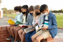 Grupo de niños multiétnicos que usan máscaras para leer libros mientras están sentados en la pared durante un descanso. Educación primaria distanciamiento social seguridad sanitaria durante la pandemia del Coronavirus Covid19. - foto de stock