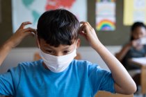 Хлопчик змішаної раси в масці для обличчя, сидячи на своєму столі в школі. Початкова освіта соціальне дистанціювання охорона здоров'я під час пандемії коронавірусу Covid19 . — стокове фото