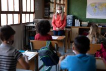 Profesora caucásica con máscara facial enseñando en clase en la escuela. Educación primaria distanciamiento social seguridad sanitaria durante la pandemia del Coronavirus Covid19. - foto de stock