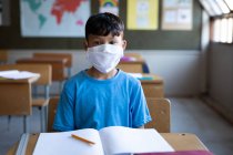 Un garçon de race mixte portant un masque facial assis sur son bureau à l'école. Enseignement primaire distanciation sociale sécurité sanitaire pendant la pandémie de coronavirus Covid19. — Photo de stock
