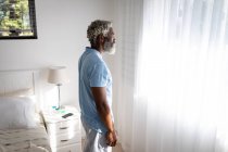 Uomo anziano afroamericano in piedi in una camera da letto, guardando attraverso una finestra, distanza sociale e isolamento in quarantena — Foto stock