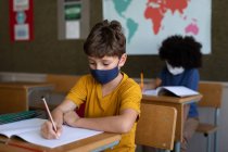 Dos niños multiétnicos sentados en escritorios con máscaras faciales en el aula. Educación primaria distanciamiento social seguridad sanitaria durante la pandemia del Coronavirus Covid19. - foto de stock