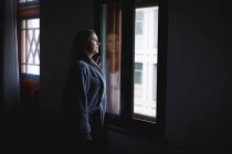 Кавказская женщина с длинными темными волосами наслаждается временем дома, социальной дистанцированностью и самоизоляцией в карантинной изоляции, стоя и глядя в окно. — стоковое фото