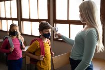 Кавказька вчителька, одягнена в маску обличчя, вимірює температуру дітей в початковій школі. Первинна освіта Соціальна безпека для здоров'я під час пандемії Ковіда19 Коронавірус. — стокове фото