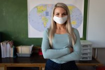 Портрет кавказької вчительки з масками на обличчі стоїть зі схрещеними в класі руками. Первинна освіта Соціальна безпека для здоров'я під час пандемії Ковіда19 Коронавірус — стокове фото
