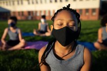 Chica de raza mixta con máscara facial realizando yoga en el jardín de la escuela. Educación primaria distanciamiento social seguridad sanitaria durante la pandemia del Coronavirus Covid19. - foto de stock