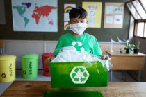 Retrato de un chico de raza mixta con máscara facial sosteniendo un contenedor de reciclaje en clase en la escuela. Educación primaria distanciamiento social seguridad sanitaria durante la pandemia del Coronavirus Covid19. - foto de stock
