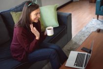 Mulher caucasiana desfrutando de tempo em casa, distanciamento social e auto-isolamento em quarentena, sentada no sofá na sala de estar, usando um laptop, acenando durante videochamada. — Fotografia de Stock