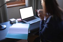 Femme caucasienne profitant du temps à la maison, de la distance sociale et de l'isolement personnel en quarantaine, assise à table, utilisant un ordinateur portable, buvant du café. — Photo de stock