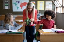 Eine kaukasische Lehrerin zeigt einer Gruppe multiethnischer Kinder in der Schule einen Pflanztopf. Grundschulbildung soziale Distanzierung der Gesundheitssicherheit während der Covid19 Coronavirus-Pandemie. — Stockfoto
