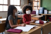 Multi ethnic boy and girl sitting at desks wearing face masks in classroom. Primary education social distancing health safety during Covid19 Coronavirus pandemic. — Stock Photo