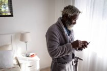 Афроамериканський старший чоловік стоїть у спальні, користуючись смартфоном, соціальною дистанцією та самоізоляцією в карантинному блокуванні. — стокове фото