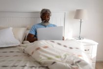 Afro-americano idoso deitado em uma cama em um quarto, usando um laptop e sorrindo, distanciamento social e auto-isolamento em quarentena bloqueio — Fotografia de Stock