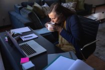 Femme caucasienne profitant du temps à la maison, de la distance sociale et de l'isolement personnel en quarantaine, assise à table, utilisant un ordinateur portable et buvant du thé. — Photo de stock